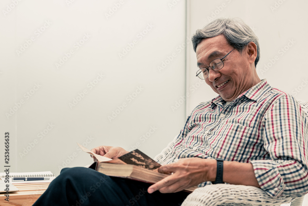 Happy senior Asian man reading book on the chair in living room at home. Retirement lifestyle and ag