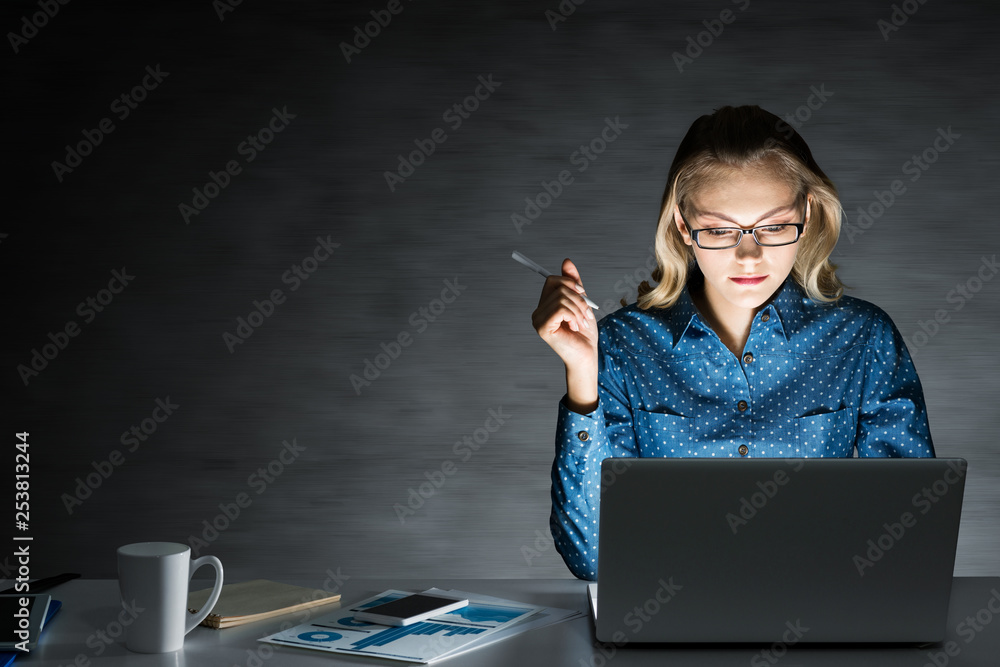 Attractive blonde working on laptop in dark office. Mixed media