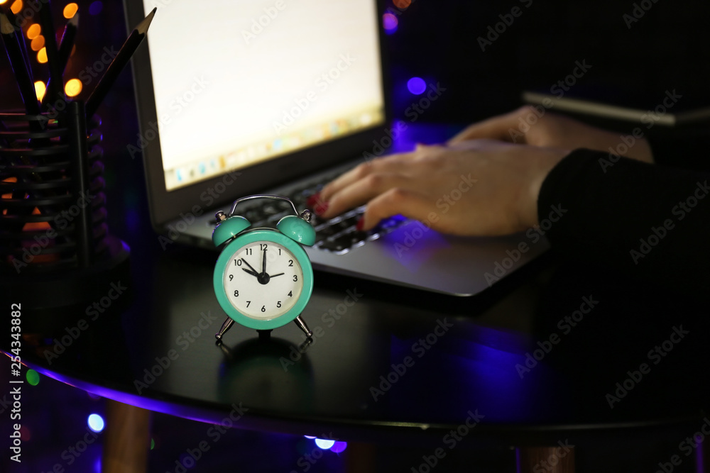 Alarm clock on table of woman working with laptop in evening