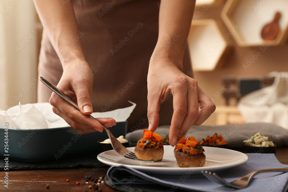 Woman putting tasty stuffed mushrooms from dish on plate in kitchen