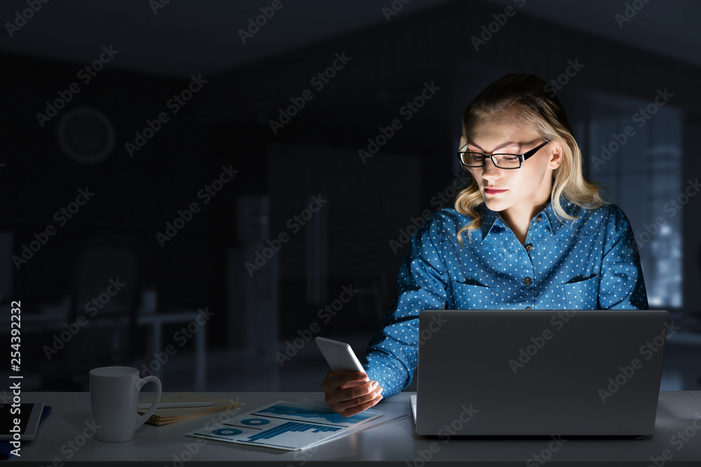 Attractive blonde working on laptop in dark office. Mixed media