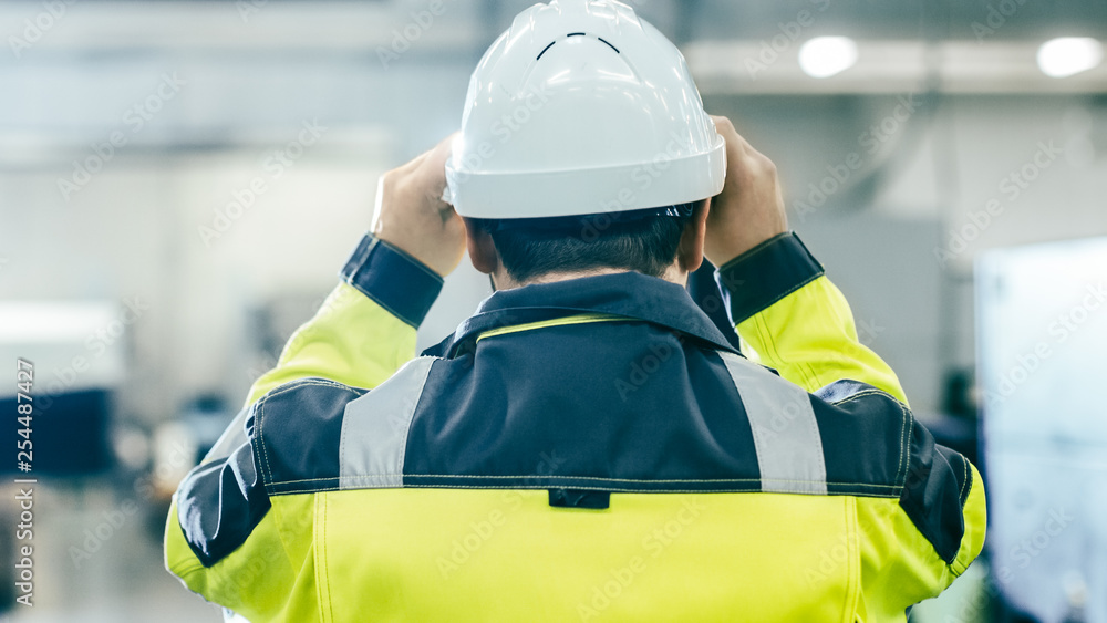 Back View Shot of the Industrial Engineer Wearing Protective Clothing Puts on Hard Hat and Walks Thr