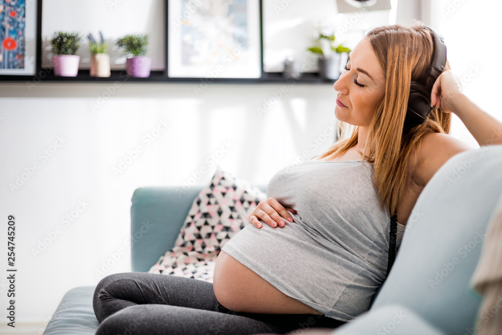 Relaxed pregnant woman sitting on sofa at home and listening music in headphones