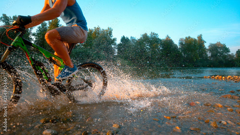 CLOSE UP: Athletic guy splashes the glassy water while cycling along a stream.