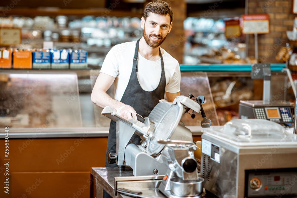 Portrait of a handsome worker in uniform slicing cheese with cutting machine in the supermarket