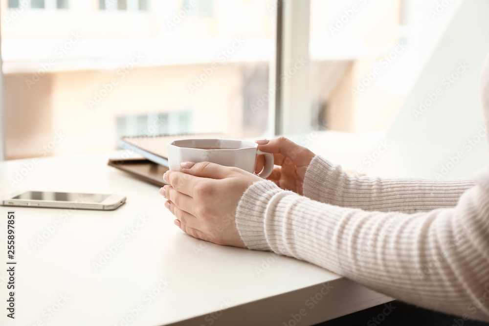 Young woman with cup of hot tea sitting at table near window at home