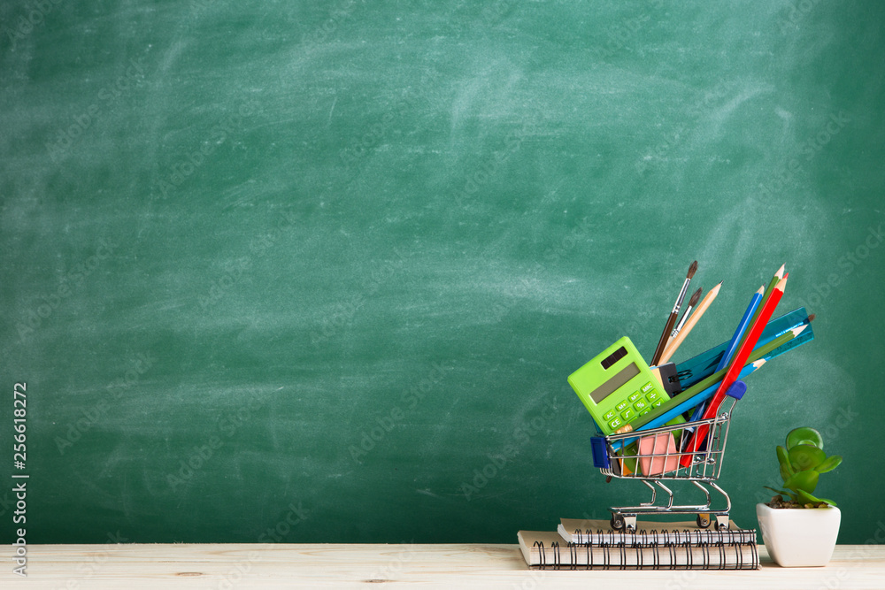 Education concept - school supplies in a shopping cart on the desk in the auditorium, blackboard bac