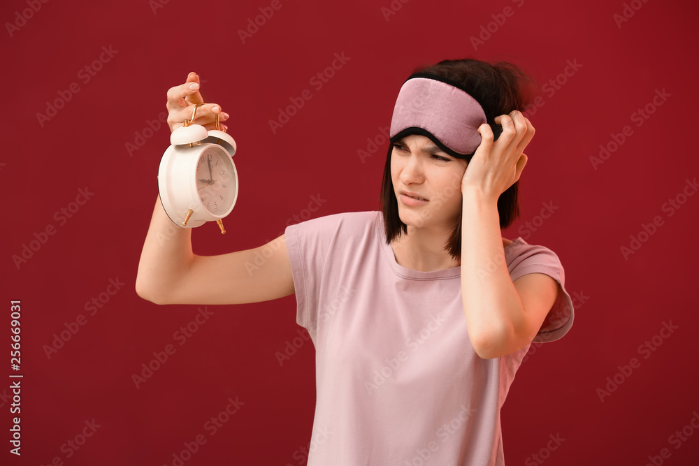 Displeased young woman with sleep mask and alarm clock on color background