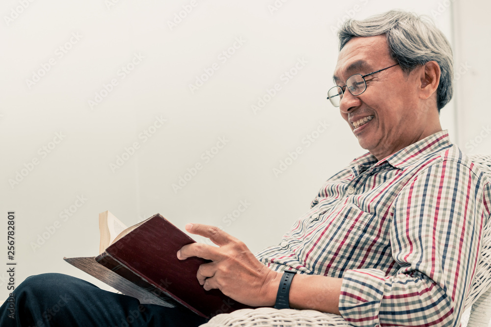 Happy senior Asian man reading book on the chair in living room at home. Retirement lifestyle and ag