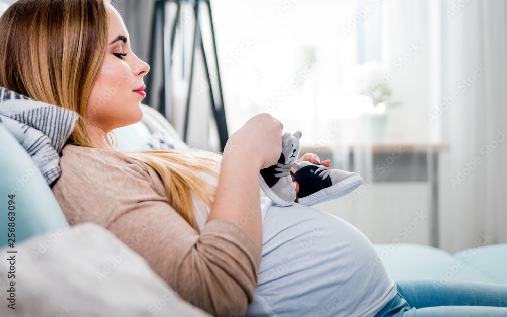 Young pregnant woman with small baby shoes sitting on sofa at home, child expectation concept