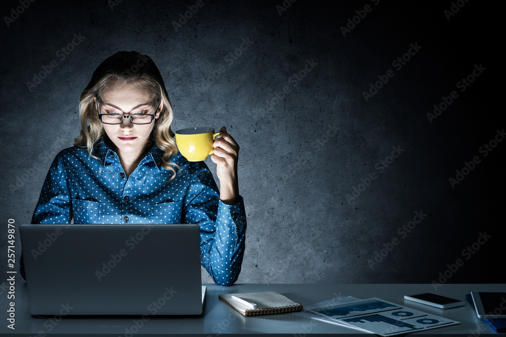 Attractive blonde working on laptop in dark office. Mixed media