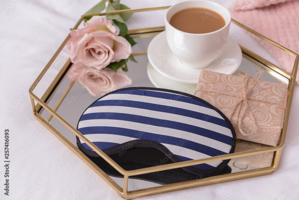 Tray with cup of coffee, flower, gift and sleep mask on bed