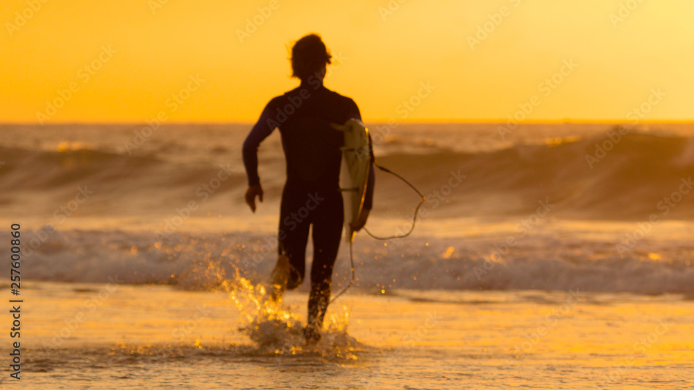 SILHOUETTE: Surfer dude holding his surfboard while running towards the ocean.