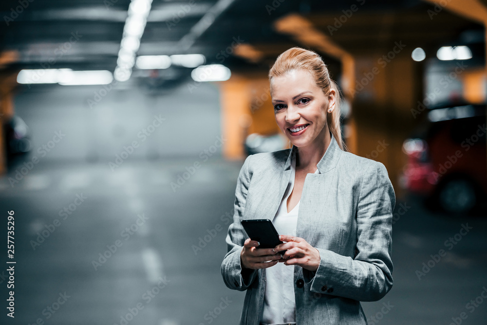 Portrait of successful businesswoman holding smartphone and looking at camera in underground car par