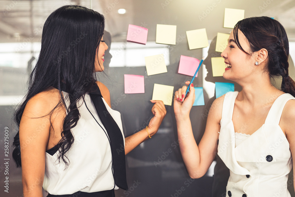 Two young beautiful Asian businesswomen talk and discuss creative post on the window. It is project 