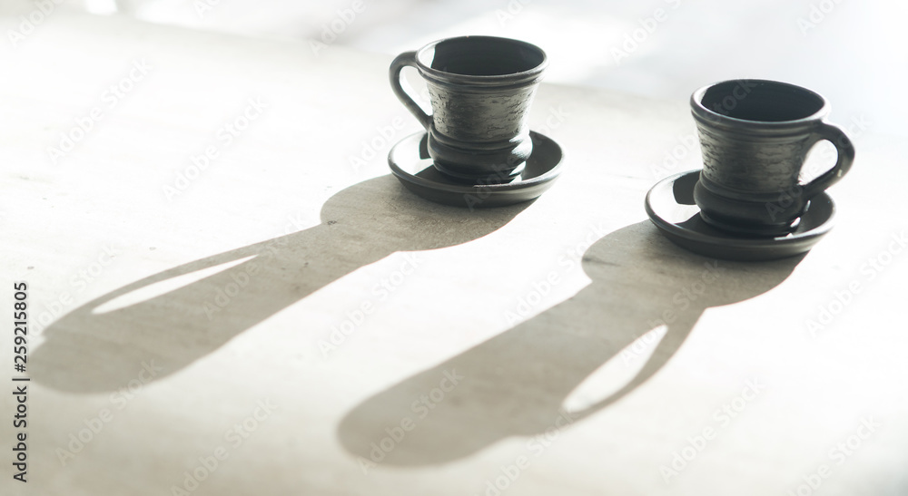 Two Coffee clay black cups on table with bright backlight which draws long cup shadows. Coffee for c