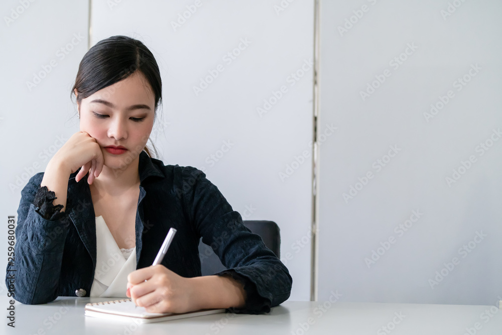 Beautiful young Asian woman writes letter on notebook while sitting at office desk. Content writer a