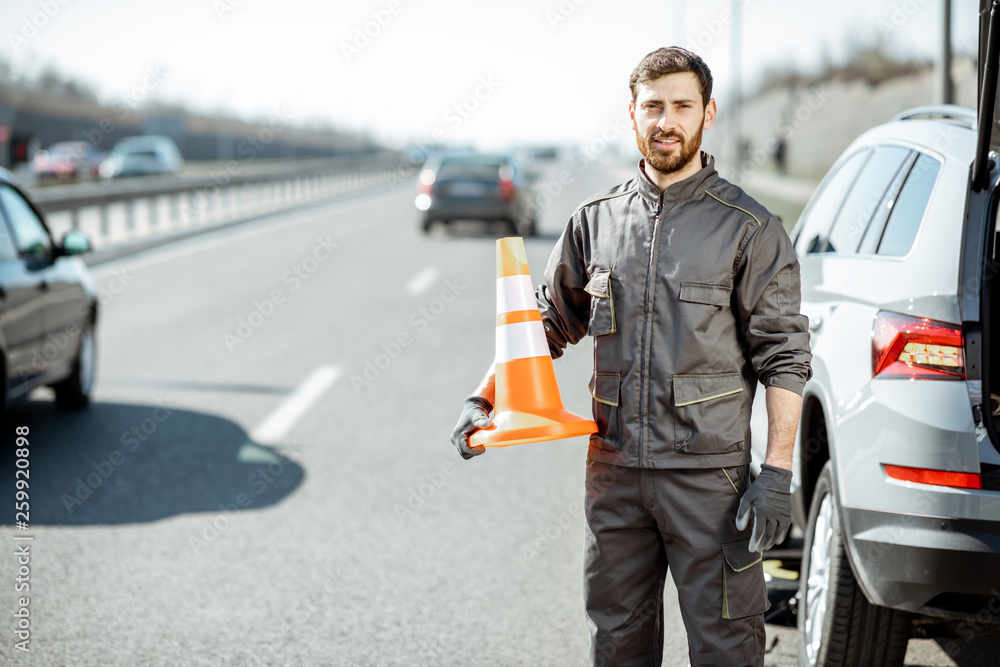 Portrait of a happy road assistance worker in uniform standing with cone near the car on the roadsid