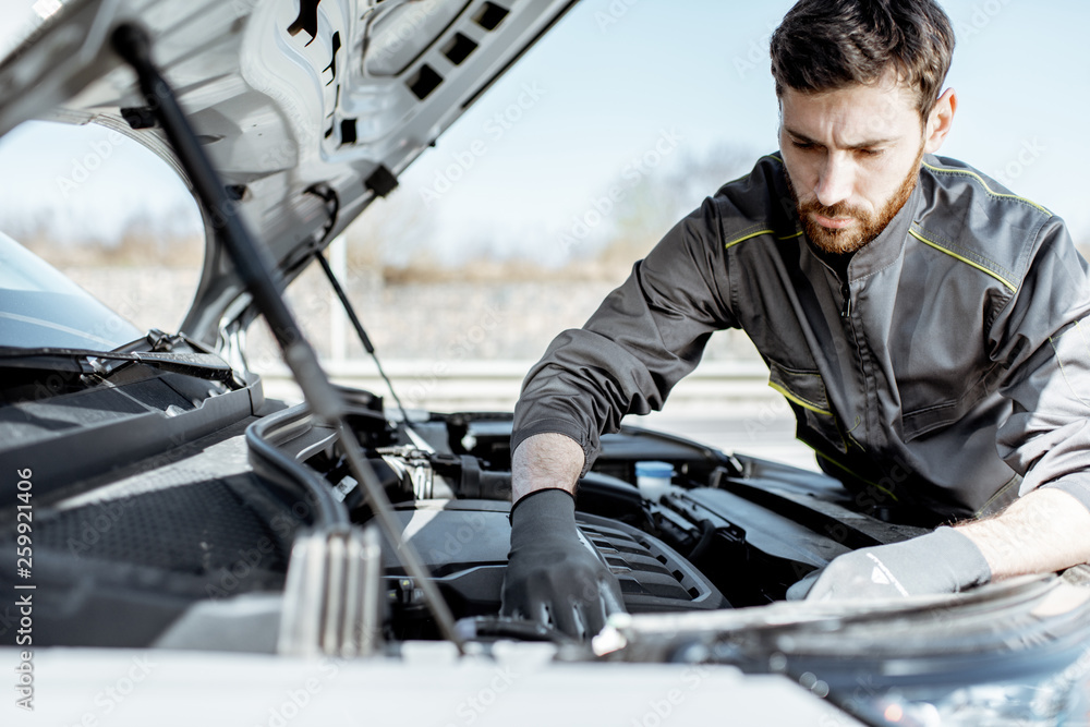 Handsome auto mechanic or road assistance worker in uniform repairing engine of the broken car on th