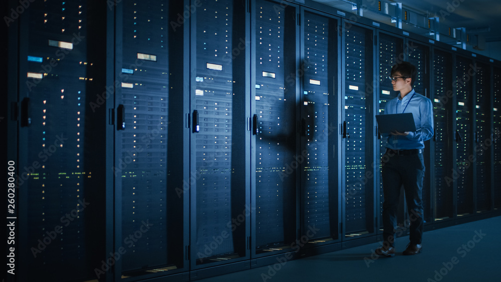 In Dark Data Center: Male IT Specialist Walks along the Row of Operational Server Racks, Uses Laptop