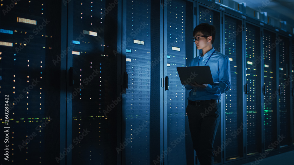 In Dark Data Center: Male IT Specialist Walks along the Row of Operational Server Racks, Uses Laptop