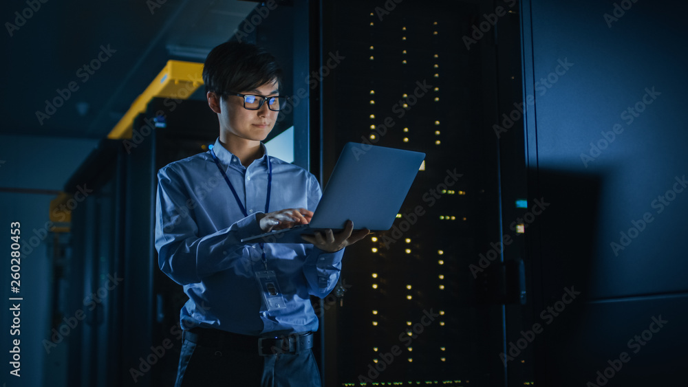 In Dark Data Center: Male IT Specialist Stands Beside the Row of Operational Server Racks, Uses Lapt