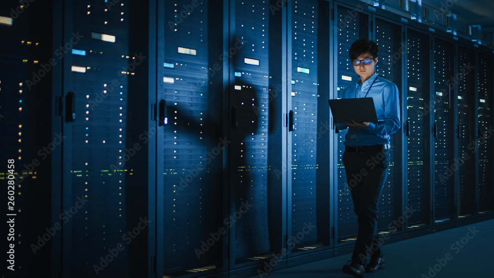 In Dark Data Center: Male IT Specialist Walks along the Row of Operational Server Racks, Uses Laptop