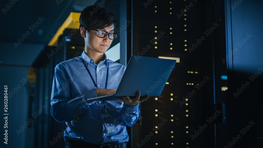 In Dark Data Center: Male IT Specialist Stands Beside the Row of Operational Server Racks, Uses Lapt