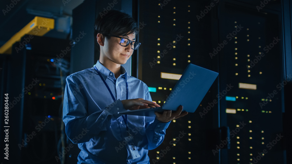 In Dark Data Center: Male IT Specialist Stands Beside the Row of Operational Server Racks, Uses Lapt