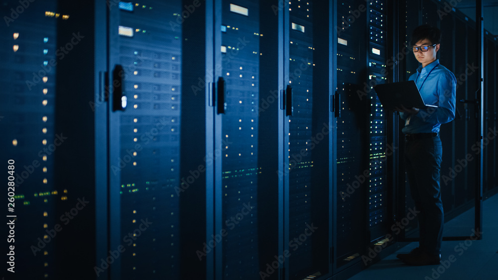 In Dark Data Center: Male IT Specialist Stands Beside the Row of Operational Server Racks, Uses Lapt