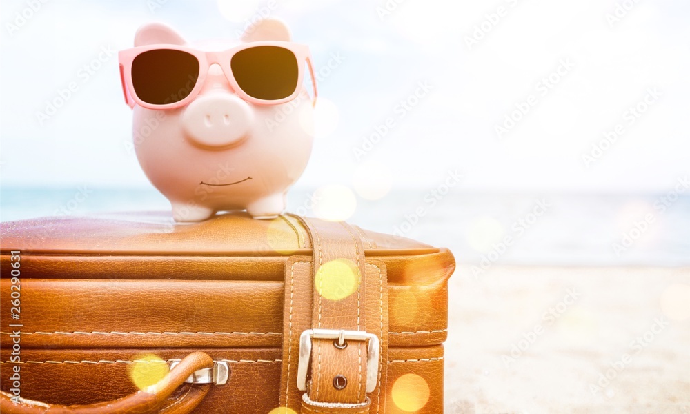 Pink piggy bank in sunglasses on leather travel case over sea beach background. Savings for vacation