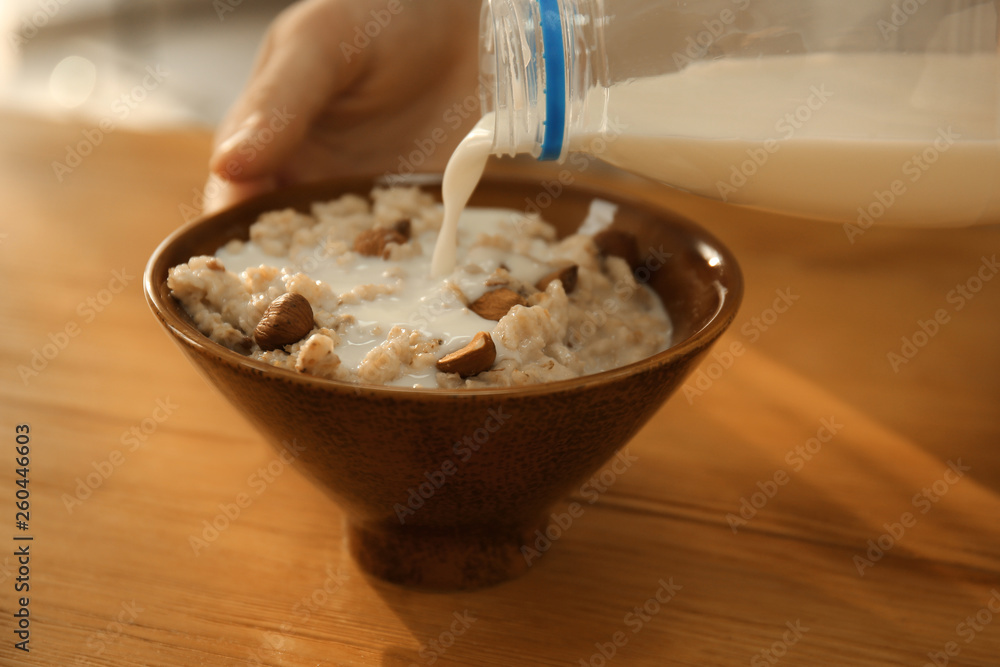 Woman pouring milk from bottle onto oatmeal in bowl on table