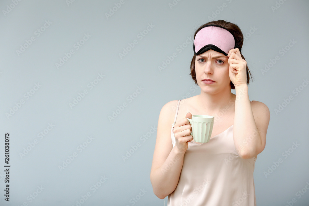 Sad woman with sleep mask and cup of coffee on color background