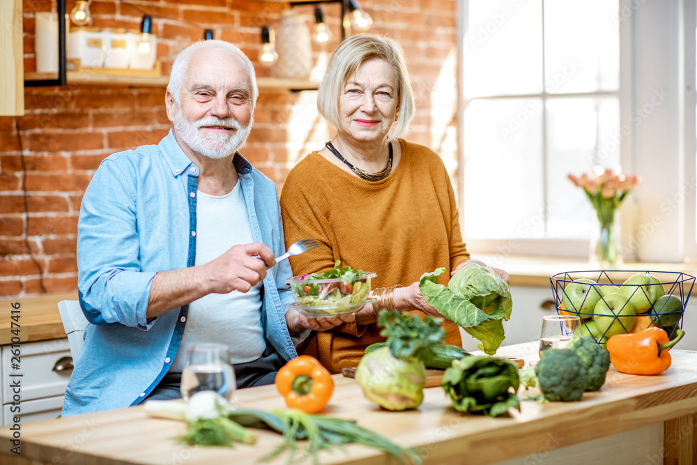 Cheerful senior couple eating salad standing together with healthy food on the kitchen at home. Conc