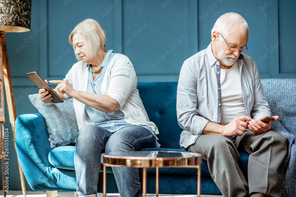 Offended senior man and woman sitting back to each other, using digital gadgets on the couch at home