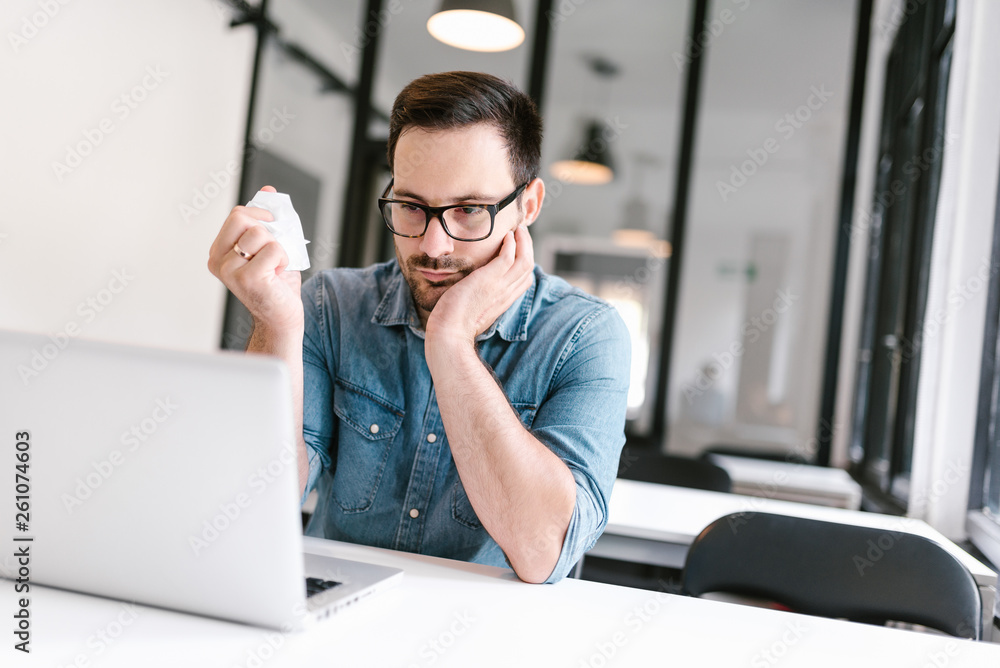 Annoyed casual man working in office while crumpling paper and looking at laptop.