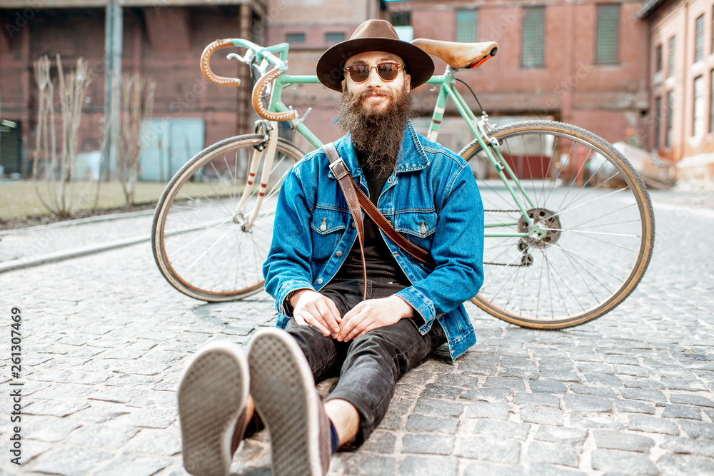 Portrait of a bearded hipster dressed stylishly with hat and jacket sitting with retro bicycle on th