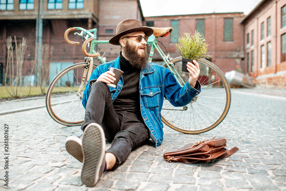 Portrait of a stylish hipster with flower pot sitting near his retro bicycle on the street on the ur