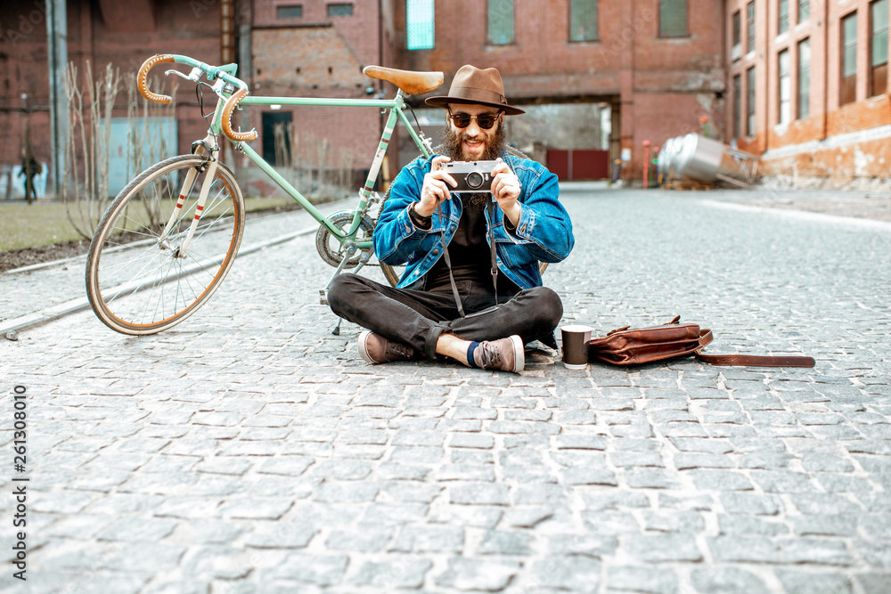 Bearded hipster dressed stylishly with hat and jacket sitting with photo camera and retro bicycle on