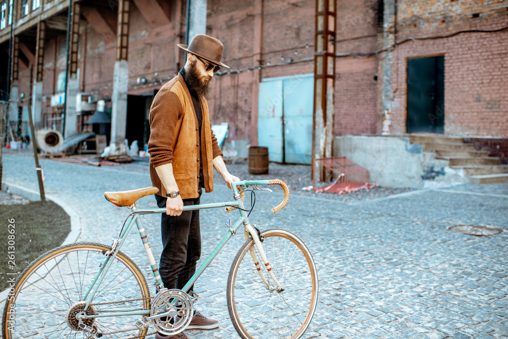 Lifestyle portrait of a bearded hipster dressed stylishly walking with retro bicycle on the industri