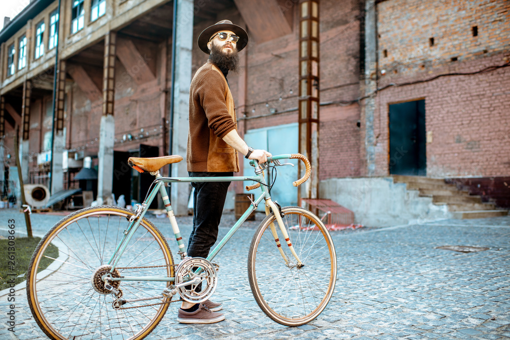 Lifestyle portrait of a bearded hipster dressed stylishly walking with retro bicycle on the industri