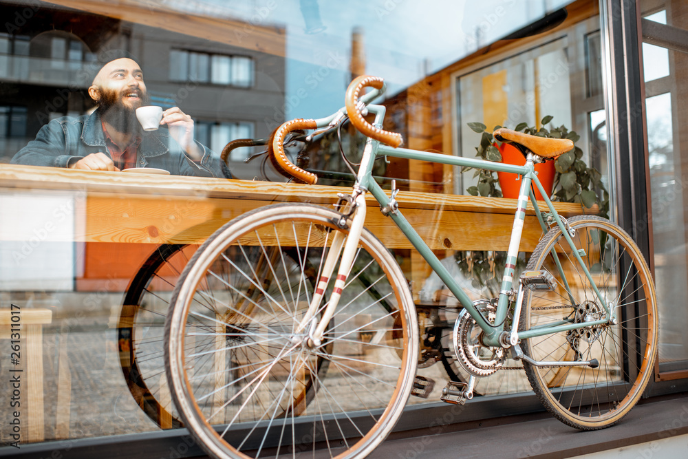 Stylish man enjoying coffee drink while sitting at the cafe near the window with retro bicycle. Wide