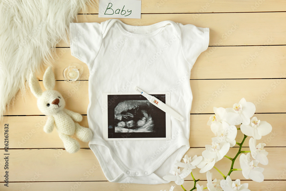 Baby clothes, pregnancy test, toy and flowers on wooden background