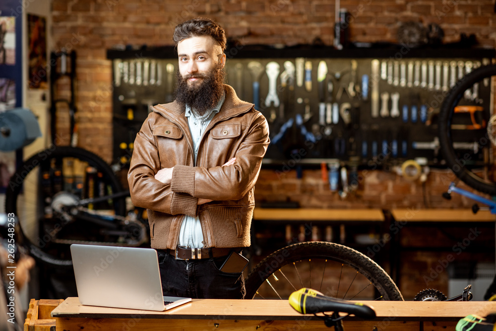 Portrait of a handsome bearded man as bicycle store owner or manager standing with laptop at the bic