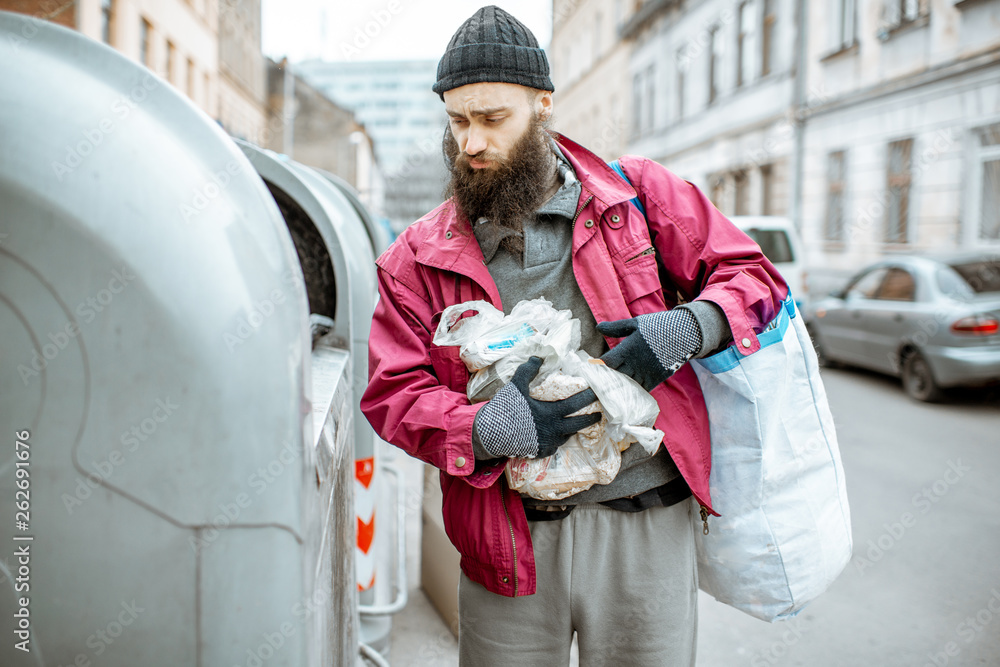 Portrait of a homeless bearded beggar standing with bag and some finded food near the trash containe