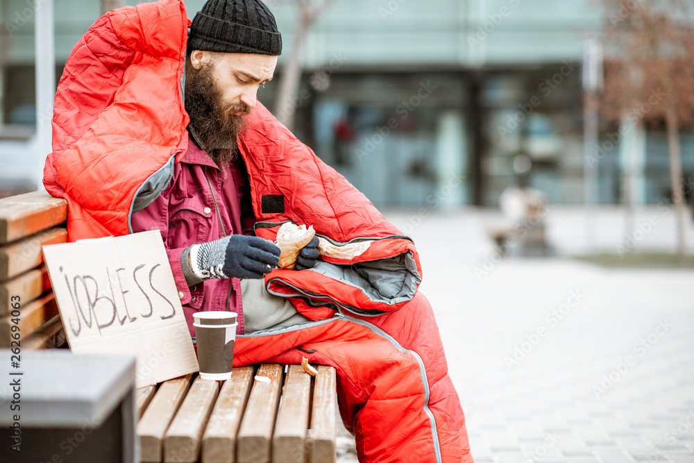 Depressed homeless beggar eating bread while sitting wrapped with sleeping bag on the bench near the