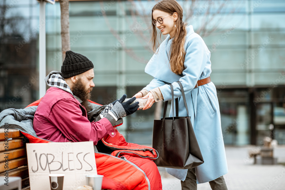 Smiling woman helping homeless beggar giving some food outdoors. Concept of helping poor people