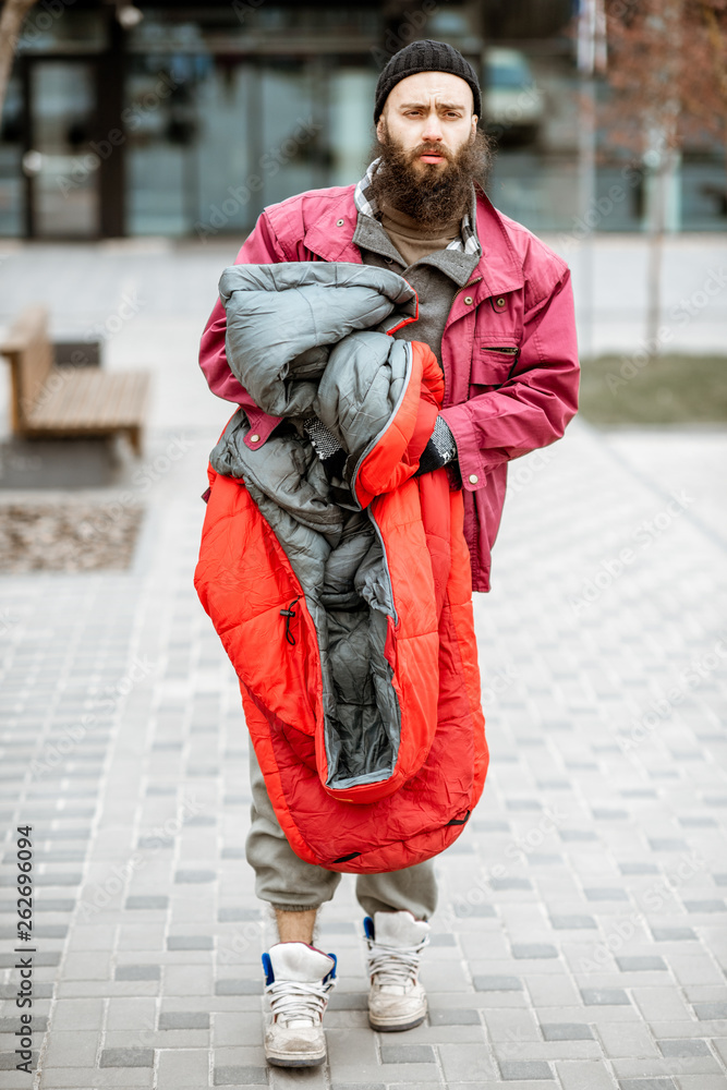 Portrait of a depressed and homeless beggar standing with sleeping bag near the business center outd