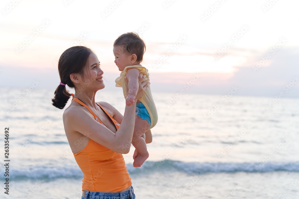 Asian mother standing on the beach holding her baby in two arms raising the kid and looking at her c