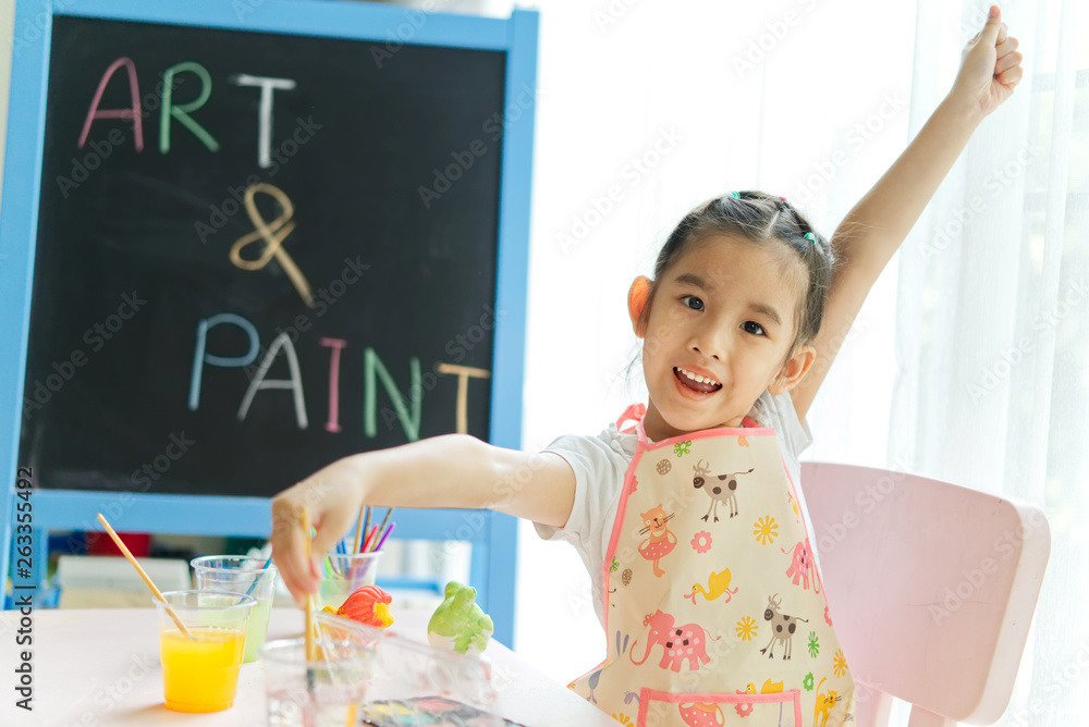 Young little Asian girl painting plaster dolls in painting class at home. She is raising the hand an